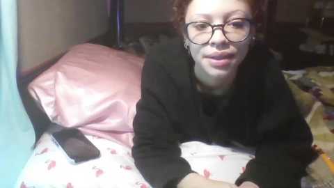 Video of a woman with fair skin and curly red hair, wearing glasses and a black top, lying on a pink pillow in a cluttered bedroom.