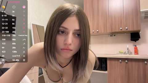 A video of a young woman with light skin and straight, shoulder-length brown hair, wearing a beige top, in a modern kitchen with wooden cabinets.