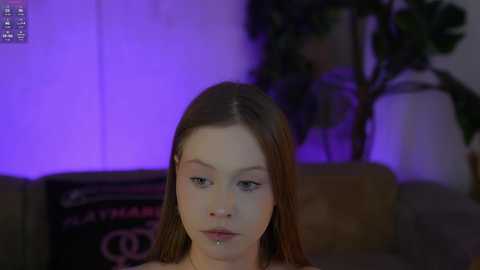 Video of a young woman with long brown hair, light skin, and a small facial piercing, sitting on a couch, with purple lighting, blurred background, and a plant to the right.