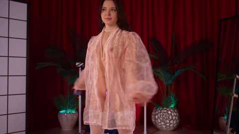 Video of a woman with medium skin tone and dark hair, wearing a sheer, pale pink robe, standing in front of red curtains with green plants and a partition in the background.
