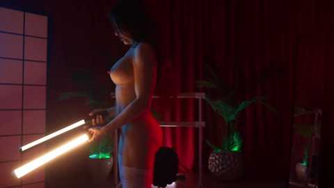 Video of a nude woman with medium skin tone, dark hair, and medium-sized breasts, standing in a dimly lit room with red curtains, green plants, and a paper lantern.