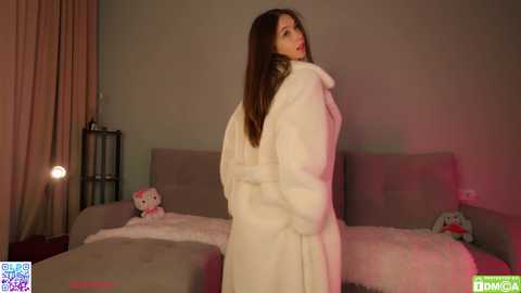 A video of a young woman with long brown hair, wrapped in a white bathrobe, standing in a dimly lit bedroom with plush grey furniture and two stuffed animals.