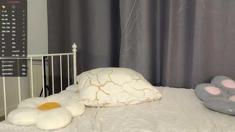 A video of a child's room featuring a white metal bed with a fluffy white pillow and a large, soft, white pillow with yellow crack-like designs. Gray curtains and a white wall with a calendar.