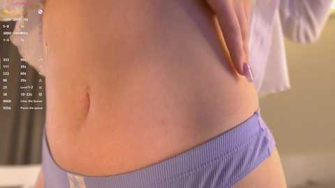 Video of a fair-skinned woman's torso, wearing light blue ribbed panties, with a hint of a belly button. Background shows a blurry indoor setting.