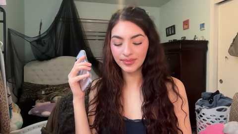 Video of a young woman with long, wavy brown hair, fair skin, and wearing a black tank top, holding a water bottle in a cluttered bedroom with a bed, clothes, and laundry basket.