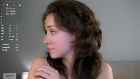 Video of a young woman with light skin and wavy brown hair, wearing a nose ring, looking contemplative, in a minimalist room with a computer screen displaying a calendar.