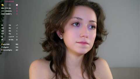 Video of a young, light-skinned woman with long, wavy brown hair, wearing small pearl earrings, looking contemplative against a plain gray background.