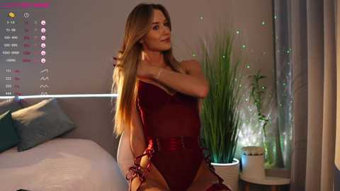 Video of a slim, long-haired woman in a deep red, sheer bodysuit, standing in a modern bedroom with a bed, plants, and a digital display on the wall.