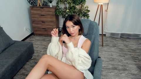 Video of a young woman with light skin and straight brown hair, wearing a white sweater, sitting on a blue armchair, petting a white cat. Background includes a grey couch, wooden dresser, potted plant, and a lamp.