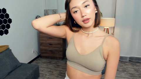 Video of a young Asian woman with straight brown hair, light skin, wearing a beige crop top and pearl necklace, standing in a modern living room with wooden furniture, hexagonal wall art, and a floor lamp.