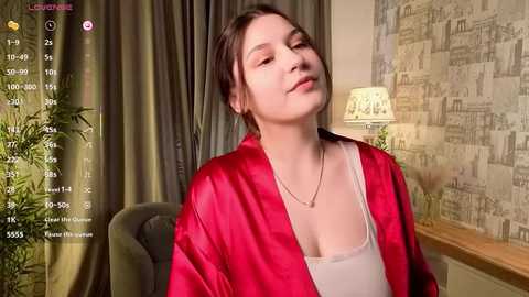 A video of a young woman with fair skin, dark hair, and a light red satin robe, sitting indoors, smiling, with a cozy, dimly lit room in the background.