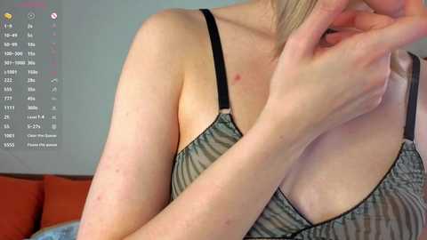 Video of a fair-skinned woman with blonde hair, wearing a striped tank top, scratching her upper arm, with a digital clock displaying time and temperature in the background.