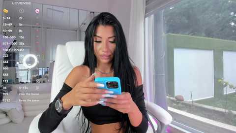 Video of a Latina woman with long black hair, wearing a black off-shoulder top, taking a selfie in a modern, minimalist room with white walls and a large window.