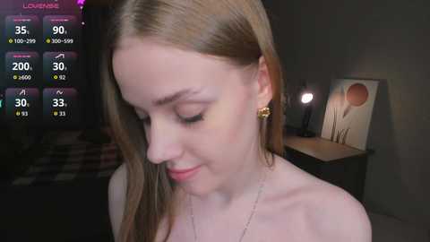 Video of a young Caucasian woman with fair skin and straight, light brown hair. She wears a gold earring and a thin necklace. Background shows a dimly lit room with a bed, a nightstand, and a wall-mounted lamp.