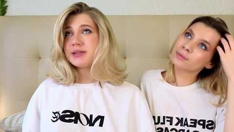 Video of two blonde women with light skin, wearing matching white t-shirts with \"LV\" logos, sitting on a beige tufted couch, one with hand in hair, the other gazing.