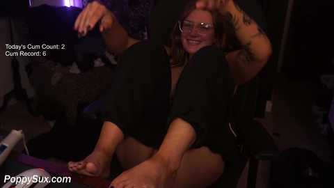 Video of a smiling, topless woman with tattoos, wearing glasses, seated on a chair in a dimly lit room, with text overlay and watermark.