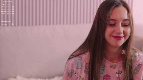 Video of a smiling, fair-skinned young woman with long brown hair, wearing pink pajamas with cartoon characters, sitting on a white couch with a white blanket. Soft, pastel-colored background.