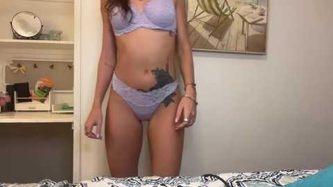 Video of a light-skinned woman in lavender lace lingerie with a tattoo on her hip, standing in a bedroom with a white wall and abstract art.