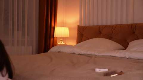 Video of a cozy, warmly lit bedroom with beige bedding, a tufted headboard, and a bedside lamp. A woman's hand holding a phone is visible on the bed.