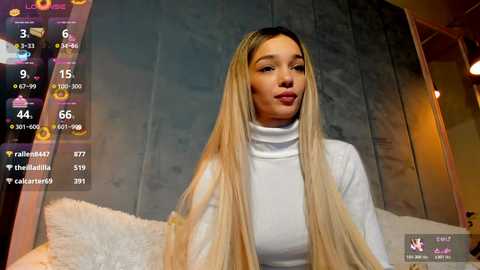 A video of a young woman with long, straight blonde hair, wearing a white turtleneck sweater, sitting indoors with a cozy, dimly lit background.