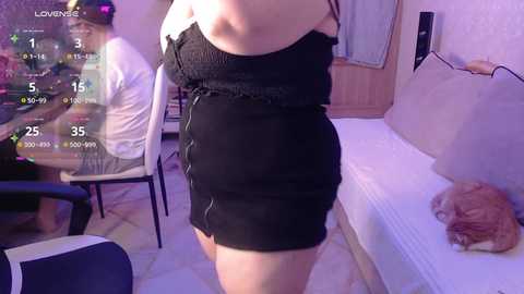 Video of a light-skinned woman with small breasts wearing a tight black dress, standing next to a beige sofa with a sleeping cat. Background includes a purple-lit room with a table, chairs, and a wooden door.