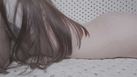 A video of a nude woman lying face down, showcasing her pale skin and long, straight brown hair cascading over her back. The background features a textured, white wall with small, black dots. The setting appears intimate and minimalistic.