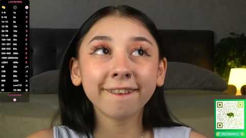 Video of a smiling young Asian girl with straight black hair, wearing a sleeveless white top, in a dimly lit bedroom. A green QR code and a digital clock with a calendar are in the background.