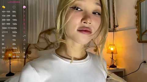 Video of a young, fair-skinned Asian woman with long, blonde hair, wearing a white shirt. Background features a warm, dimly lit room with a lamp, curtains, and framed picture.