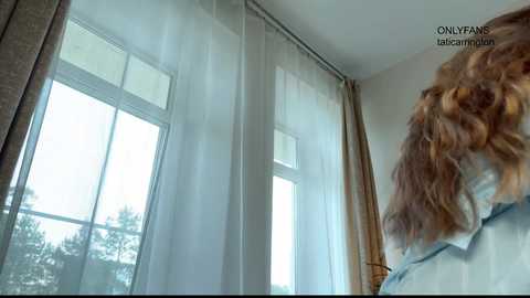A video captures a person with wavy, shoulder-length brown hair, seen from behind, standing near large windows with sheer white curtains in a brightly lit room.