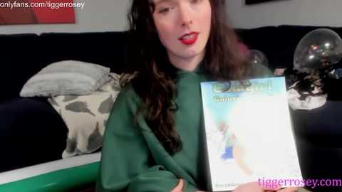 Video of a young woman with long, wavy brown hair, fair skin, and red lipstick, wearing a green hoodie, holding a book titled \"Gothic Romance.\" She sits on a dark sofa with a white blanket and a black cushion, in a cozy living room setting.