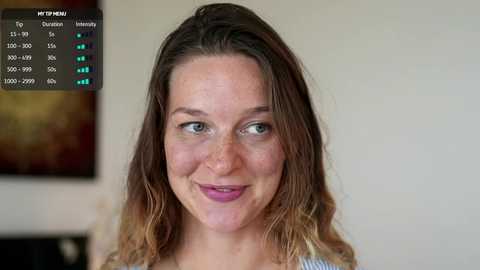 Video of a smiling Caucasian woman with light brown wavy hair, wearing a blue top, in a well-lit room with a blurred background featuring abstract art.