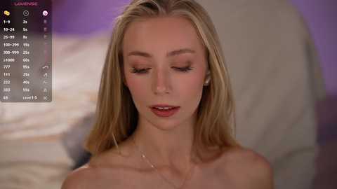 Video of a blonde, fair-skinned woman with closed eyes, wearing a necklace, on a bed with blurred purple and white sheets in the background.