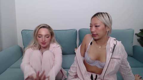 Video of two young women with light skin, one blonde and one with short platinum hair, sitting on a light blue couch. They wear matching pink robes and white lace bras, appearing relaxed and chatting.