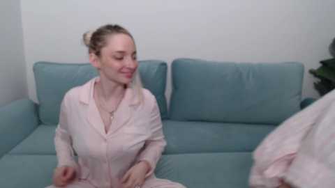 Video of a smiling, fair-skinned woman with blonde hair in a bun, wearing a light pink robe, sitting on a light blue couch in a minimalist room.