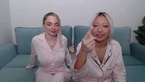 Video of two blonde women in matching pink pajamas, one gesturing with her fingers, sitting on a light blue couch in a minimalist room.