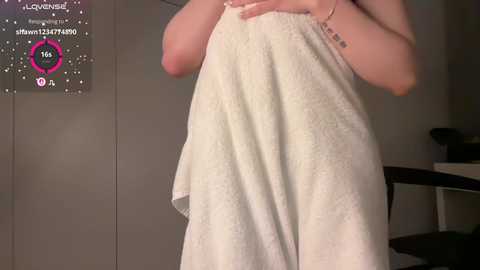 Video of a person wrapping a white towel around themselves, showing a slender physique, light skin, and a bracelet on their left wrist. Background includes a digital screen displaying a chat window and a blurry room.