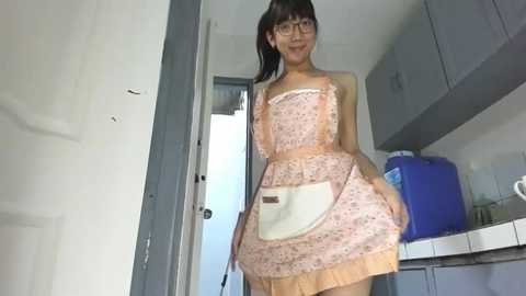 Video of a slim, young Asian woman with glasses and straight black hair, wearing a pink floral apron dress, standing in a small, simple kitchen with gray cabinets, a blue water jug, and a white sink.