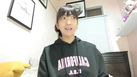 Video of a young woman with light skin and straight dark hair, wearing a black hoodie with \"Auburn\" and \"A. 1\" on it, in a cozy room with framed art, a yellow plush toy, and a white window.