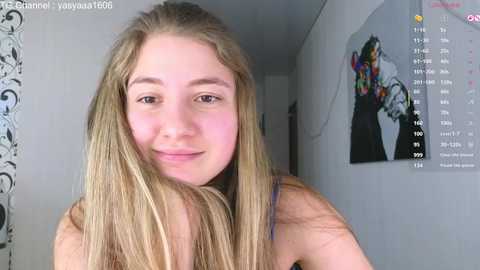 Video of a young woman with light skin, long blonde hair, and a slight smile, wearing a black top. She stands indoors, in front of a white wall with a colorful painting.