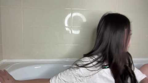 A video shows a woman with long, straight black hair in a white robe, sitting in a white bathtub, looking at her reflection in the mirror.