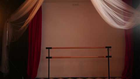 Video of a ballet barre with wooden poles and black metal supports, draped in sheer white and red curtains, set against a plain beige wall in a dimly lit studio.
