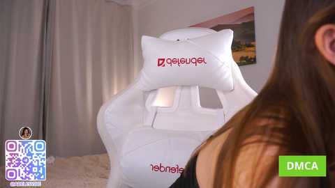 A video of a woman with long brown hair, wearing a black top, sitting on a white ergonomic gaming chair with \"geigude\" logo. The background shows a beige curtain and a yellow car picture.
