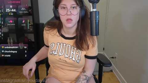 Video of a young woman with glasses, wearing a \"Dolphins\" t-shirt and headphones, sitting in a gaming chair, in a dimly-lit room with a TV and bookshelf.