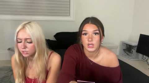 Video of two young women with long hair, one blonde, one brunette, wearing off-shoulder maroon tops, seated on a bed, with a white fan in the background.