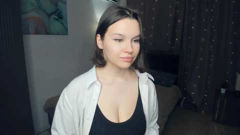 Video of a young Caucasian woman with shoulder-length dark hair, wearing a black tank top and open white shirt, standing in a dimly lit room with a bed and curtains in the background.