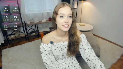 A video of a young, light-skinned woman with long, wavy brown hair, wearing a white off-shoulder dress with black floral patterns, sitting on a beige carpet in a modern, brightly lit living room.
