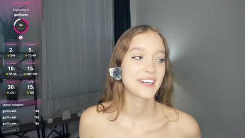 Video of a young, light-skinned woman with shoulder-length brown hair, wearing a headset, smiling in a minimalist room with a digital display showing stats.