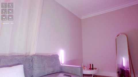 Video of a minimalist bedroom with a light gray sofa, sheer white curtains, and a tall mirror reflecting the soft purple light. A white side table holds makeup items and a small plant.