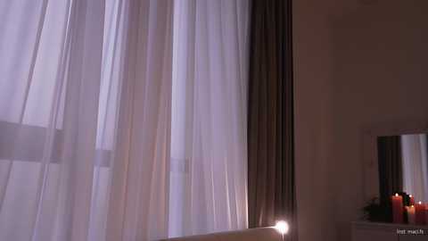 A video of a softly lit room with sheer white curtains partially drawn, revealing a glimpse of a cityscape at night. A mirror reflects the dimly lit interior, and a table holds a red candle and greenery.