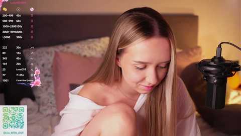 Video of a young, fair-skinned woman with long blonde hair, wearing a white off-shoulder top, sitting on a bed, with a microphone stand and recording equipment in the background.